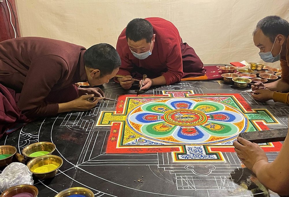 Trois moines à plat ventre font couler du sable coloré d'un tube de cuivre pour dessiner un mandala