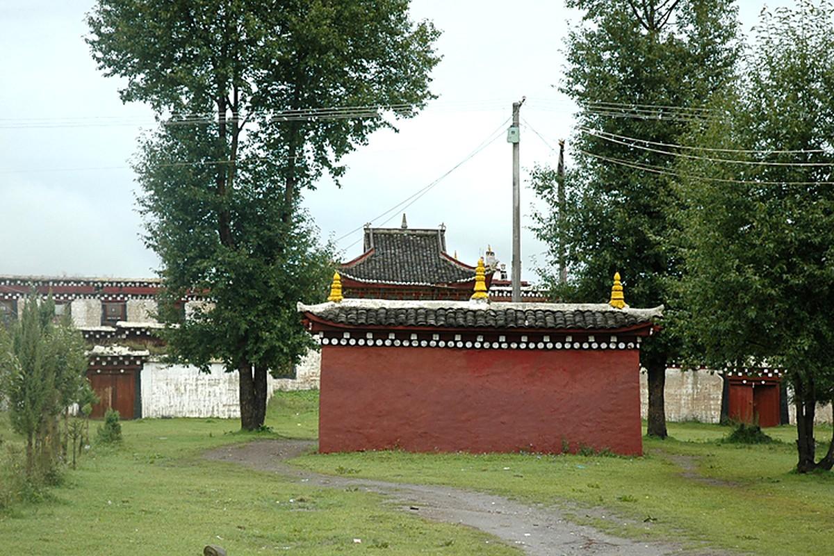 A red monastery wall with gilded finials on top, trees behind, and a barn roof beyond