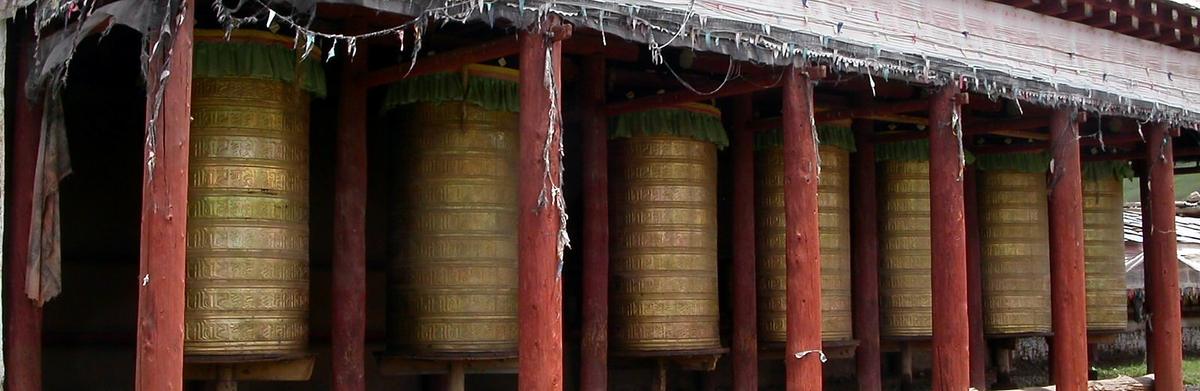 A long open corridor of large brass prayer wheels between red wooden columns, prayer flags overhead