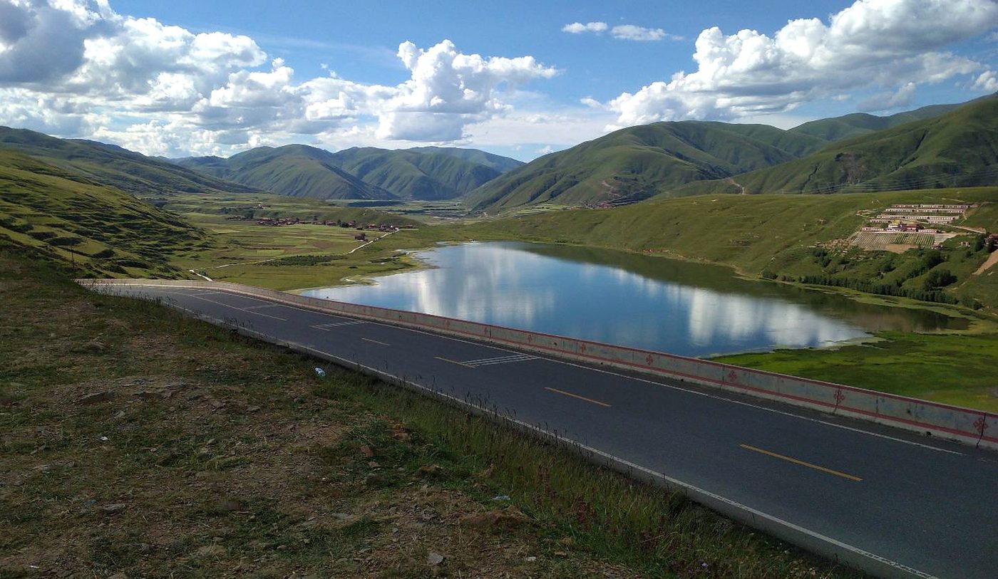 Kasa lake seen from the G317: a wide blue lake in a green basin, the road with a low barrier in the foreground, villages and terraced fields on the far slope