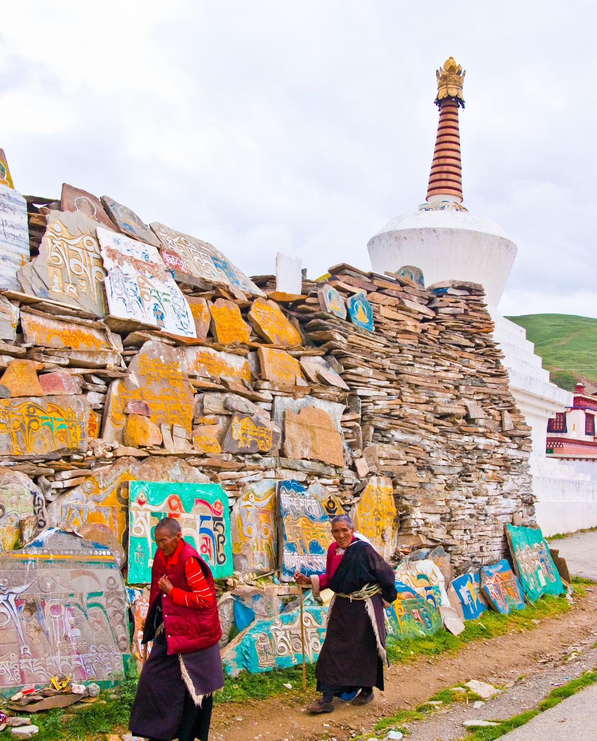 Mani slabs stacked into a wall, painted in colour, with a white stupa behind and two pilgrims walking past