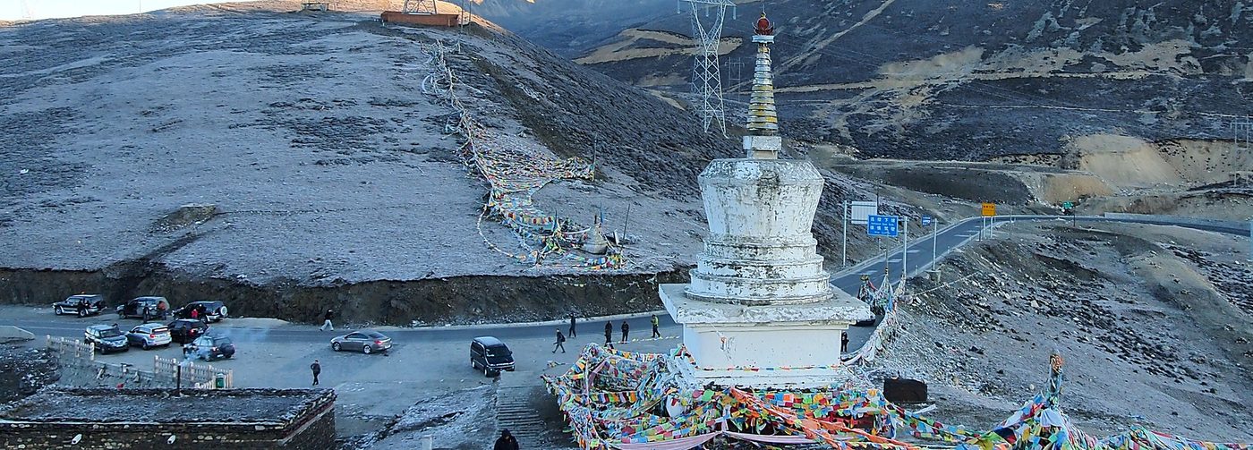 A mountain pass in western Sichuan with a white stupa, prayer flags, a car park and snow on the ground