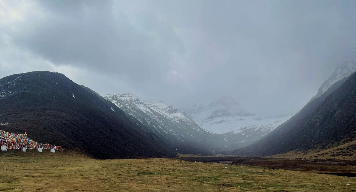 陰天下的山谷,左側坡腳掛著經幡,谷底一條河,遠處雪峰壓著雲